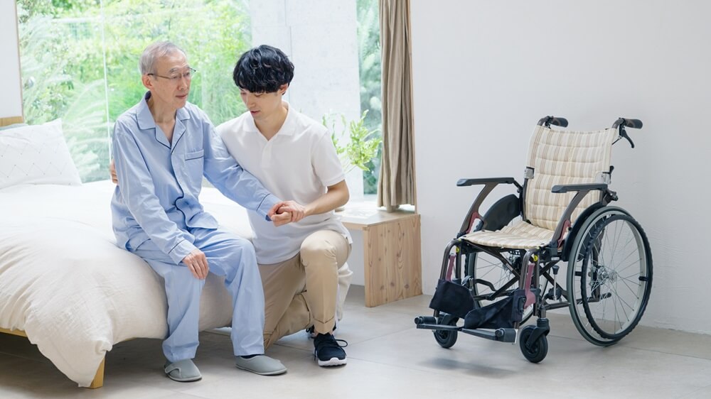 介護士がベッドのそばで高齢男性を支えている様子。近くには車椅子が置かれている。