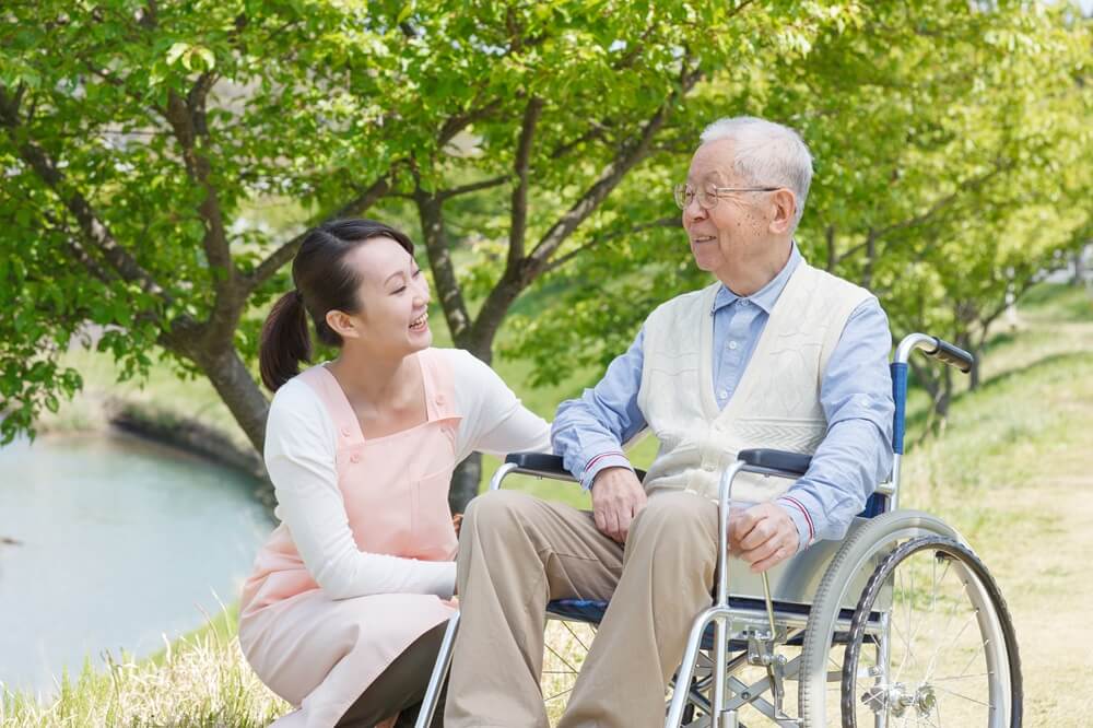 車椅子に座る高齢男性と介護士が屋外の公園で笑顔で会話している様子