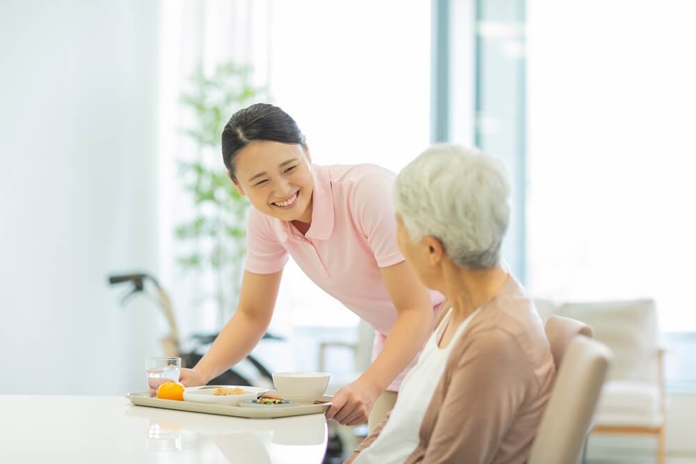 笑顔の介護士が高齢女性に食事を提供している様子。背景には車椅子が見える。